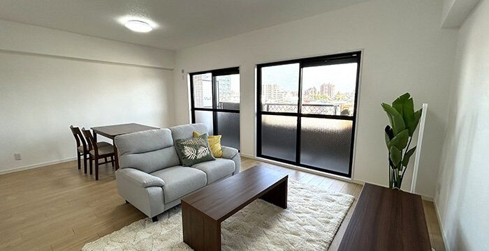 Modern living room with light wood flooring, a gray sofa, and a wooden coffee table on a white rug; sliding windows offer a city view, with a dining table and chairs in the background and a TV stand along the wall.