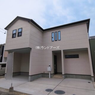 Front view of a beige, two-story modern house with an angular roof and a paved driveway; a silver van is parked to the left, with Japanese text on the facade.