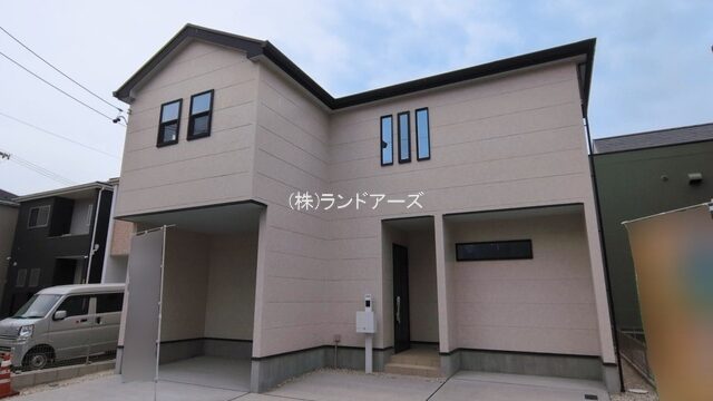 Front view of a beige, two-story modern house with an angular roof and a paved driveway; a silver van is parked to the left, with Japanese text on the facade.