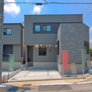 Modern two-story gray residence with a stone-clad corner, small rectangular windows, and a concrete driveway; colorful banners along the curb and a utility pole in front.