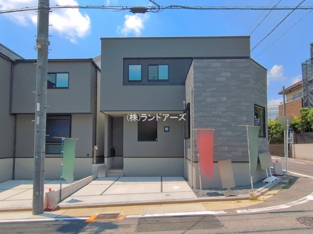 Modern two-story gray residence with a stone-clad corner, small rectangular windows, and a concrete driveway; colorful banners along the curb and a utility pole in front.