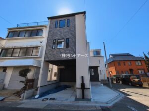 Narrow, modern three-story house with a dark brick facade on a residential street under a clear blue sky.