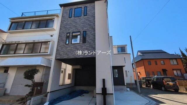 Narrow, modern three-story house with a dark brick facade on a residential street under a clear blue sky.