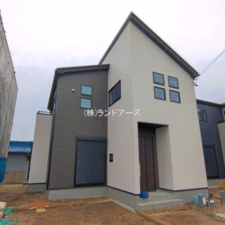Front view of a modern two-story house under construction, beige and dark gray walls, a wooden front door with steps, and construction materials in the bare yard; scaffolding on the left and a blue-tarp-covered structure nearby.