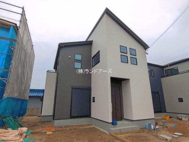 Front view of a modern two-story house under construction, beige and dark gray walls, a wooden front door with steps, and construction materials in the bare yard; scaffolding on the left and a blue-tarp-covered structure nearby.