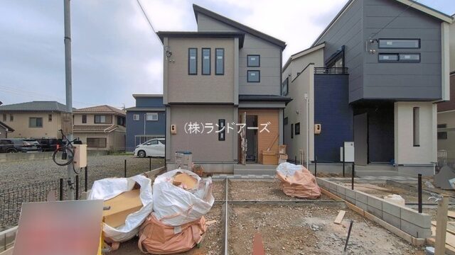 Construction site in front of two modern suburban houses; piles of wrapped construction debris on dirt lot and a traffic cone in the foreground.