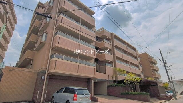 Multi-story beige apartment building with balconies along a curved street; a silver SUV parked near the entrance.