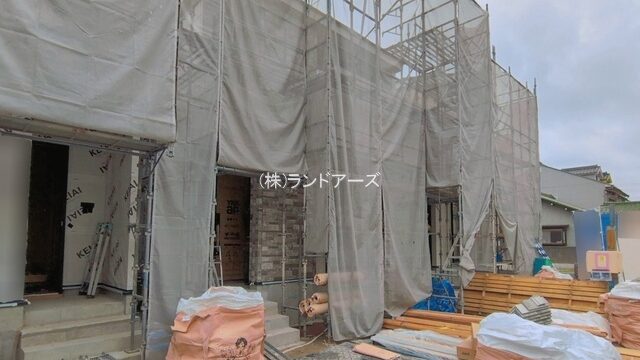 Under-construction building covered in scaffolding and tarps; construction materials and bags on the ground; company name (株)ランドアーズ overlaid.