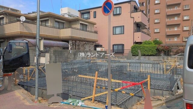 Urban construction site on a street with steel reinforcement bars, barriers, a no-parking sign, and parked vehicles near apartment buildings.