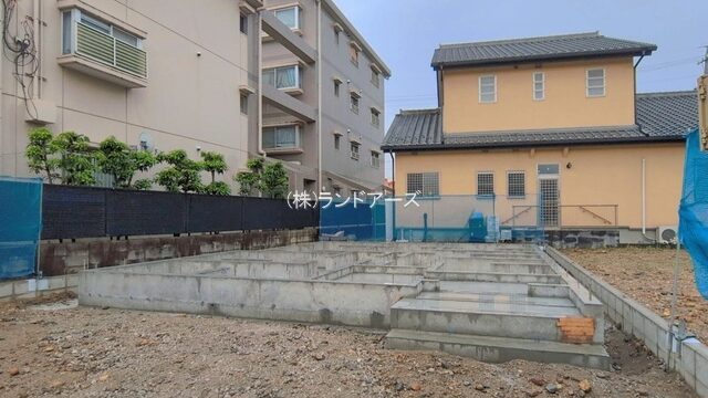 Construction site in a residential area with exposed concrete foundation and stepped platforms, apartment buildings in the background.