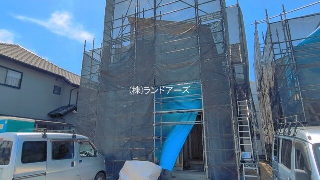 Construction site with tall scaffolding and blue netting covering a two-story building; white vans parked on either side.