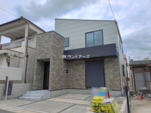 名東区猪子石11317【1】_1号棟_20260624_1_0886535 - 株式会社ランドアーズ|名古屋の一戸建て(建売・中古戸建)・中古マンション・賃貸管理・不動産売却 Modern two-story house with gray brick facade and large front window; entrance to the left and construction materials (yellow bags/tools) on the driveway.