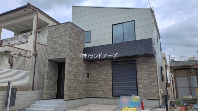 Modern two-story house with gray brick facade and large front window; entrance to the left and construction materials (yellow bags/tools) on the driveway.