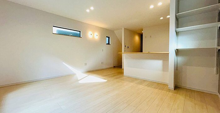 Bright empty modern living area with light wood floors, white walls, a kitchen counter, and tall open shelves on the right under ceiling lights.
