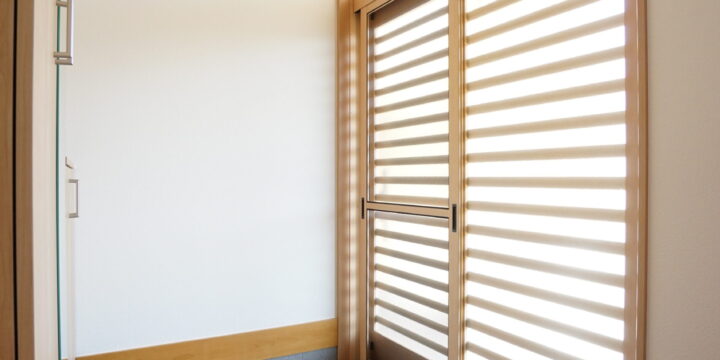 Bright entry hallway with a tall wooden-framed door featuring horizontal slatted panels and natural wood trim on white walls, letting in light across a tiled floor.