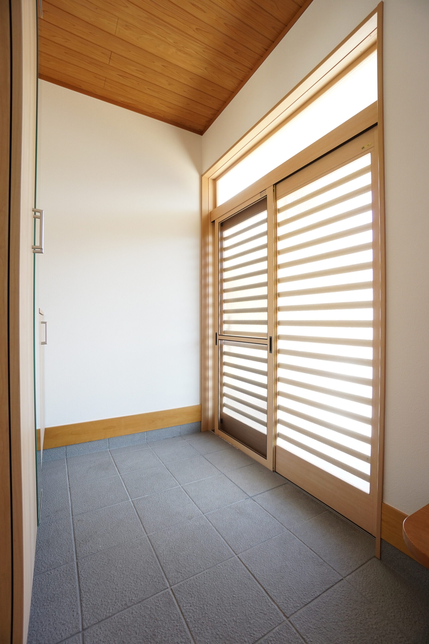 Bright entry hallway with a tall wooden-framed door featuring horizontal slatted panels and natural wood trim on white walls, letting in light across a tiled floor.