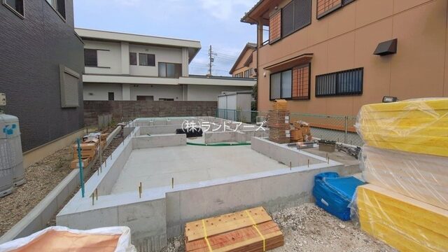 Residential construction site with concrete foundation walls being built between two houses; wooden pallets and wrapped materials on site.