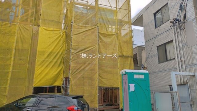 Construction site with scaffolding wrapped in yellow safety netting, a black car parked nearby, and a teal portable toilet.