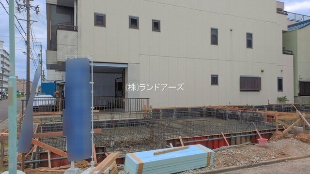 Construction site in front of a beige building, with exposed rebar and wooden formwork for a foundation, blue temporary sign, and insulation boards on the ground.